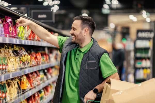 Funcionário organizando prateleira de produtos em supermercado, preparando o estoque para a Black Friday.